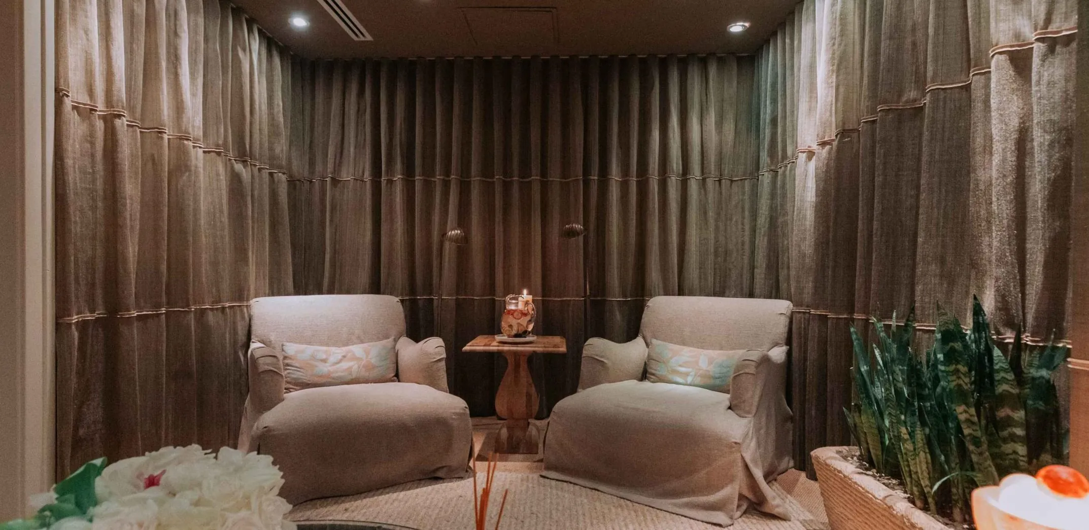 Spa room with two armchairs, draped walls, small table with candle, and soft lighting.