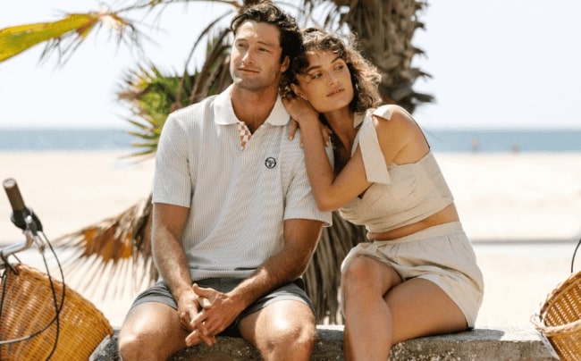 Man and woman sitting on a ledge with the ocean and palm tree in the background