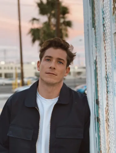 Young man with styled hair leaning against a metal fence, wearing a dark jacket over a white shirt.