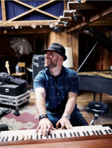 A man with a beard and hat laughing while playing a keyboard in a room with musical equipment.