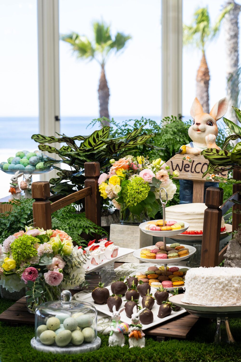 Festive table with Easter decorations, flowers, cakes, and treats, set against a beach backdrop.