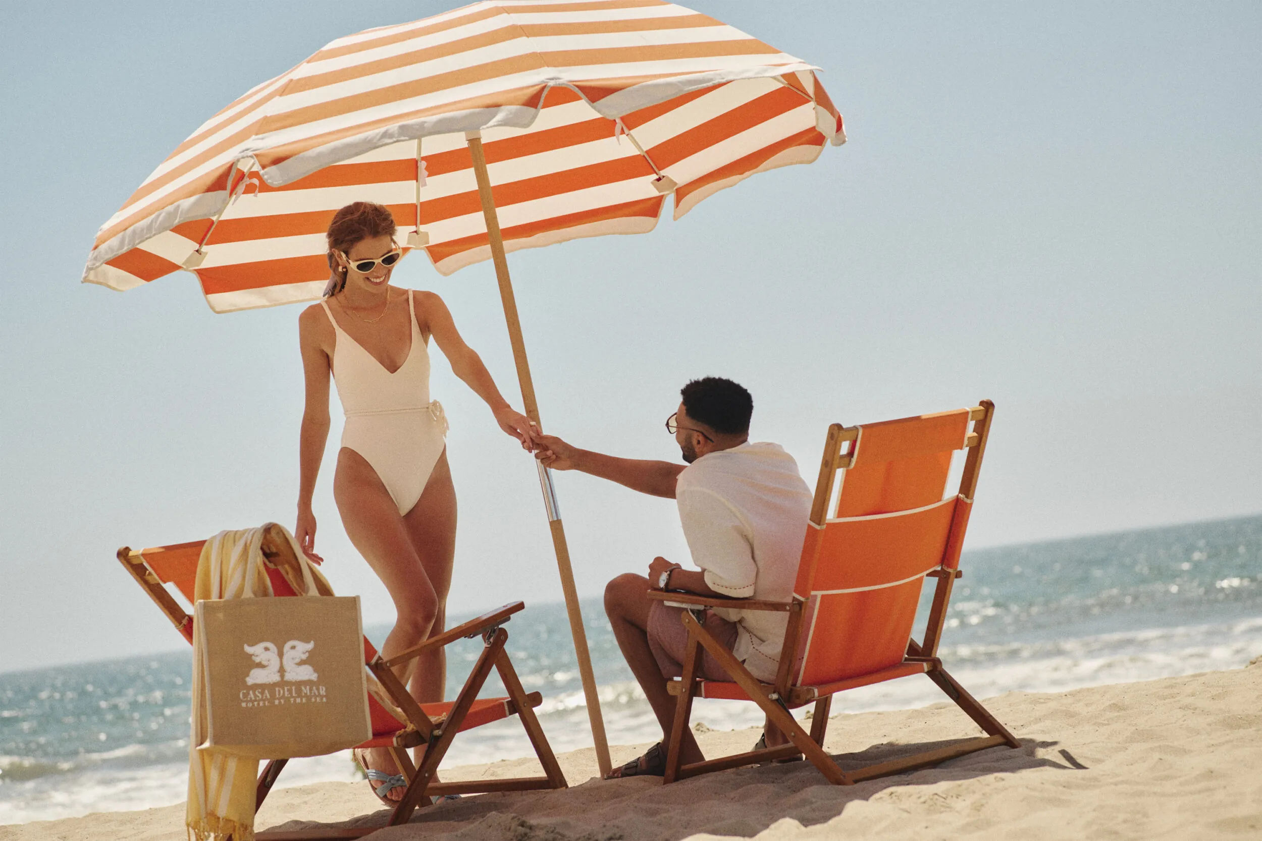 A woman holds a striped beach umbrella while a man sits on an orange beach chair in Santa Monica.