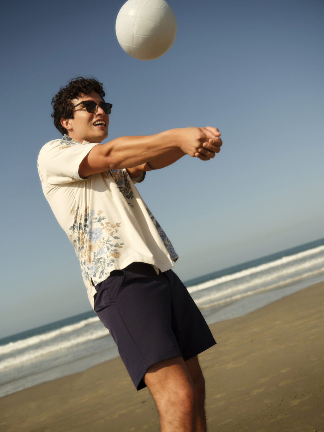 Man in sunglasses playing volleyball on a beach with a clear blue sky.