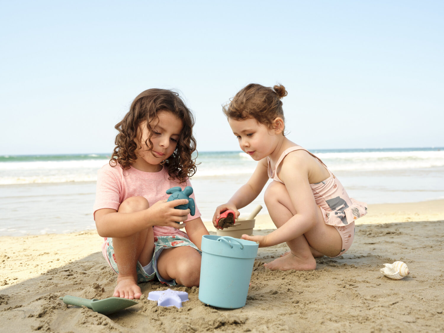 Two kids play in the sand, one holding a toy and the other with a bucket, ocean in the background.