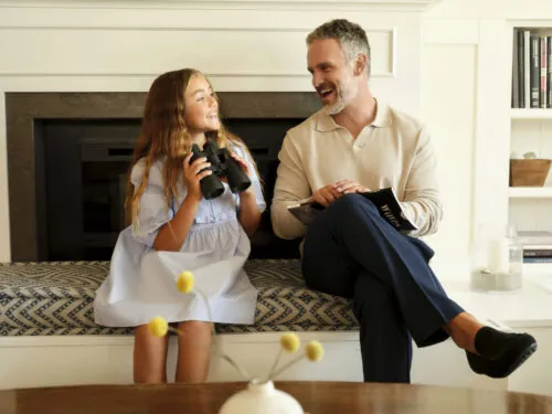 A man and a girl laughing, with the girl holding binoculars and the man holding a jacket.