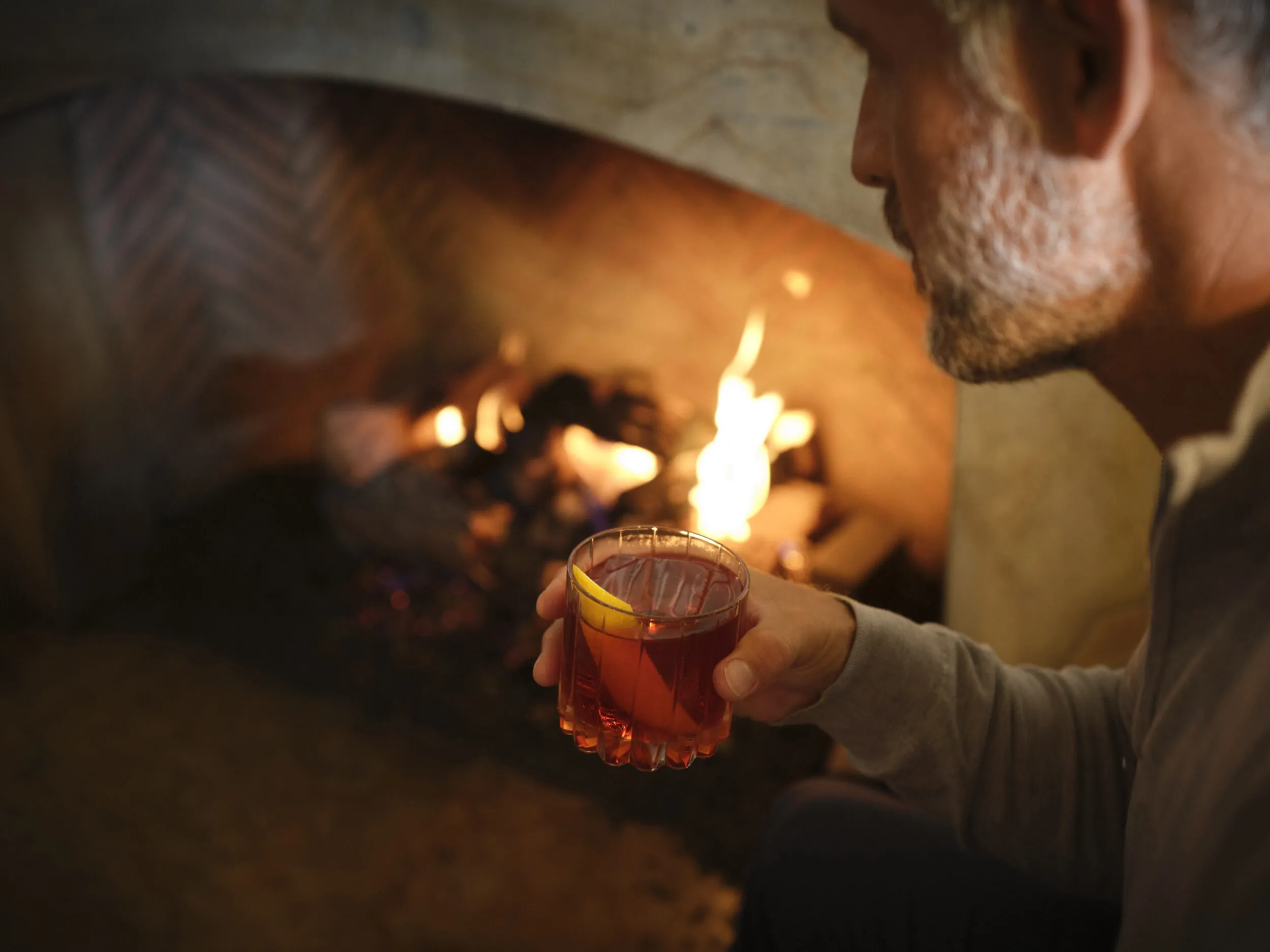 A man holding a drink with a slice of orange sits by a fireplace with visible flames.