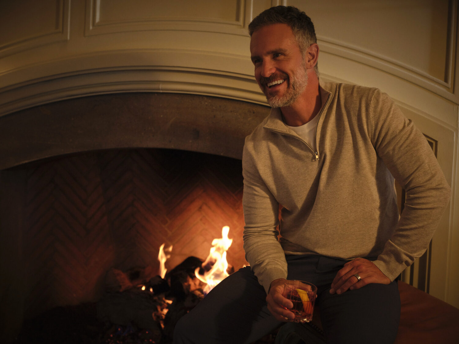 Man relaxing by the fireplace, enjoying a drink and smiling.