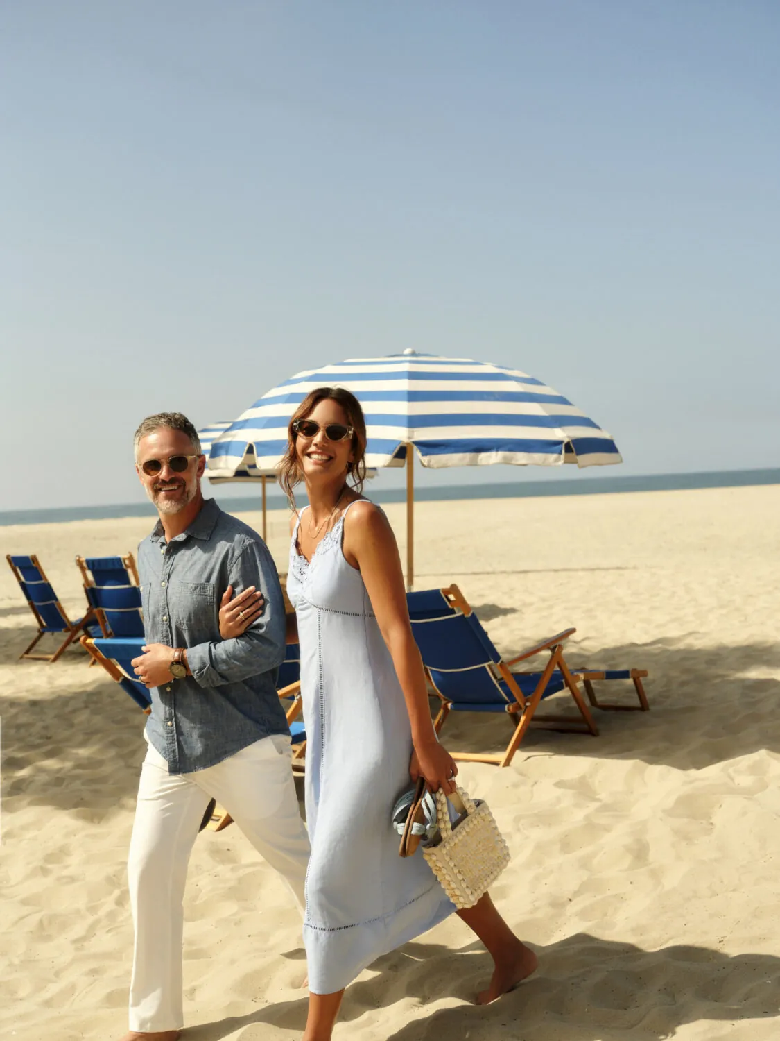 A couple walking on the beach under a striped umbrella, smiling and enjoying the sun.