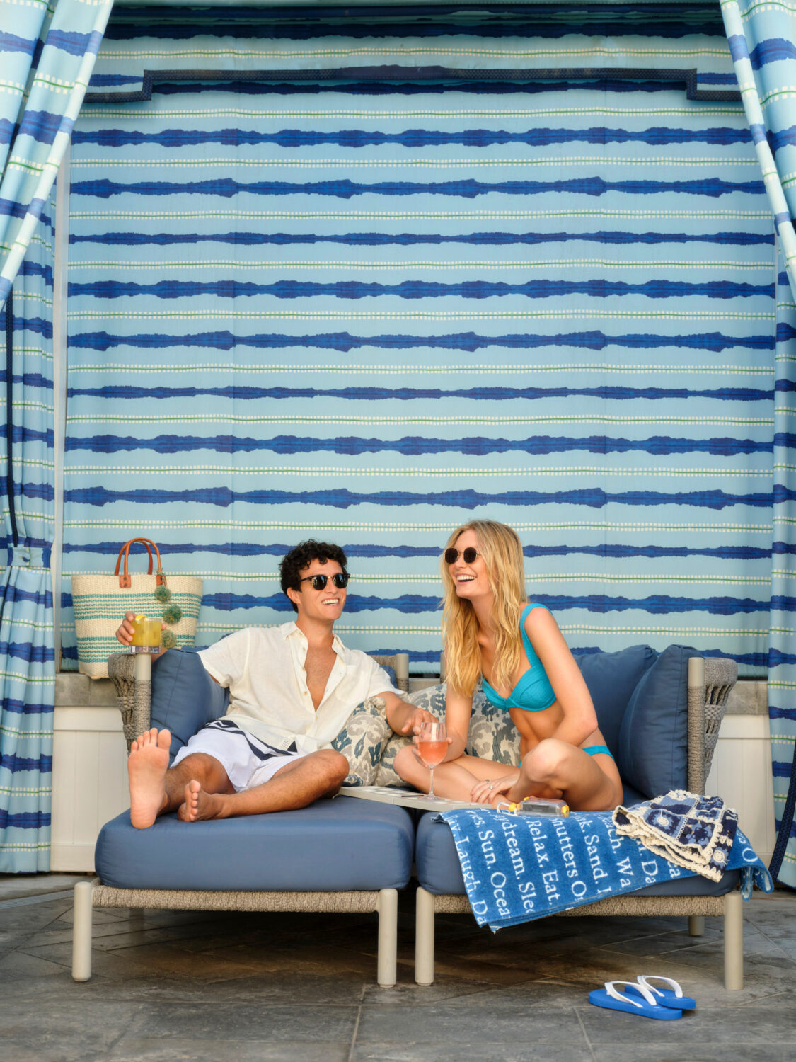Smiling couple in sunglasses relaxing in a blue cabana, enjoying drinks and conversation.