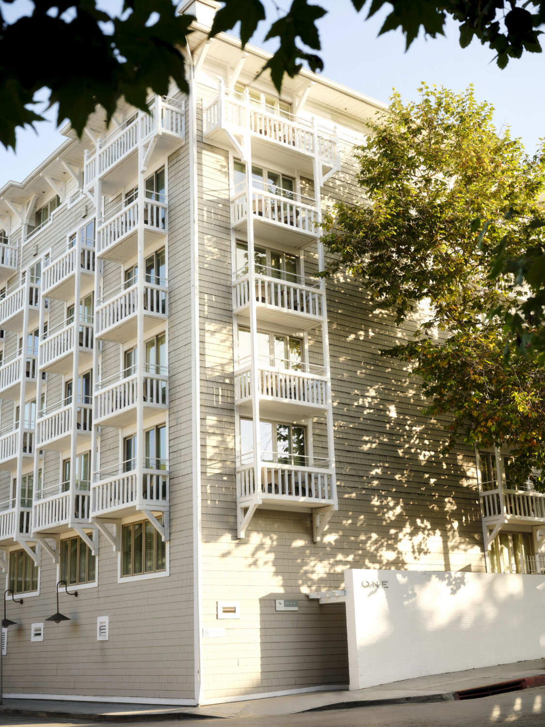 A modern building with white balconies, wooden exterior, and large windows, surrounded by greenery.