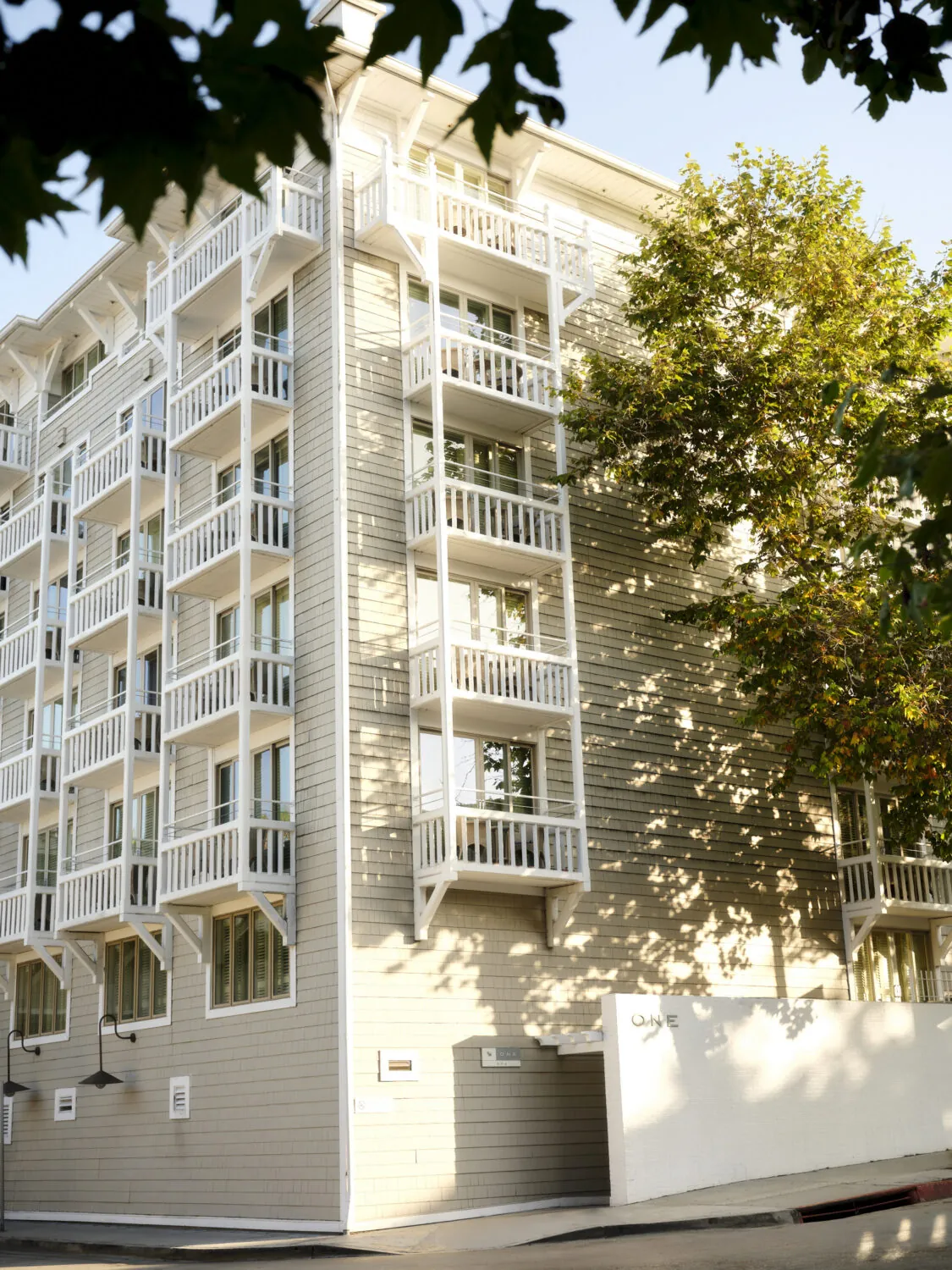 A modern building with white balconies, wooden exterior, and large windows, surrounded by greenery.