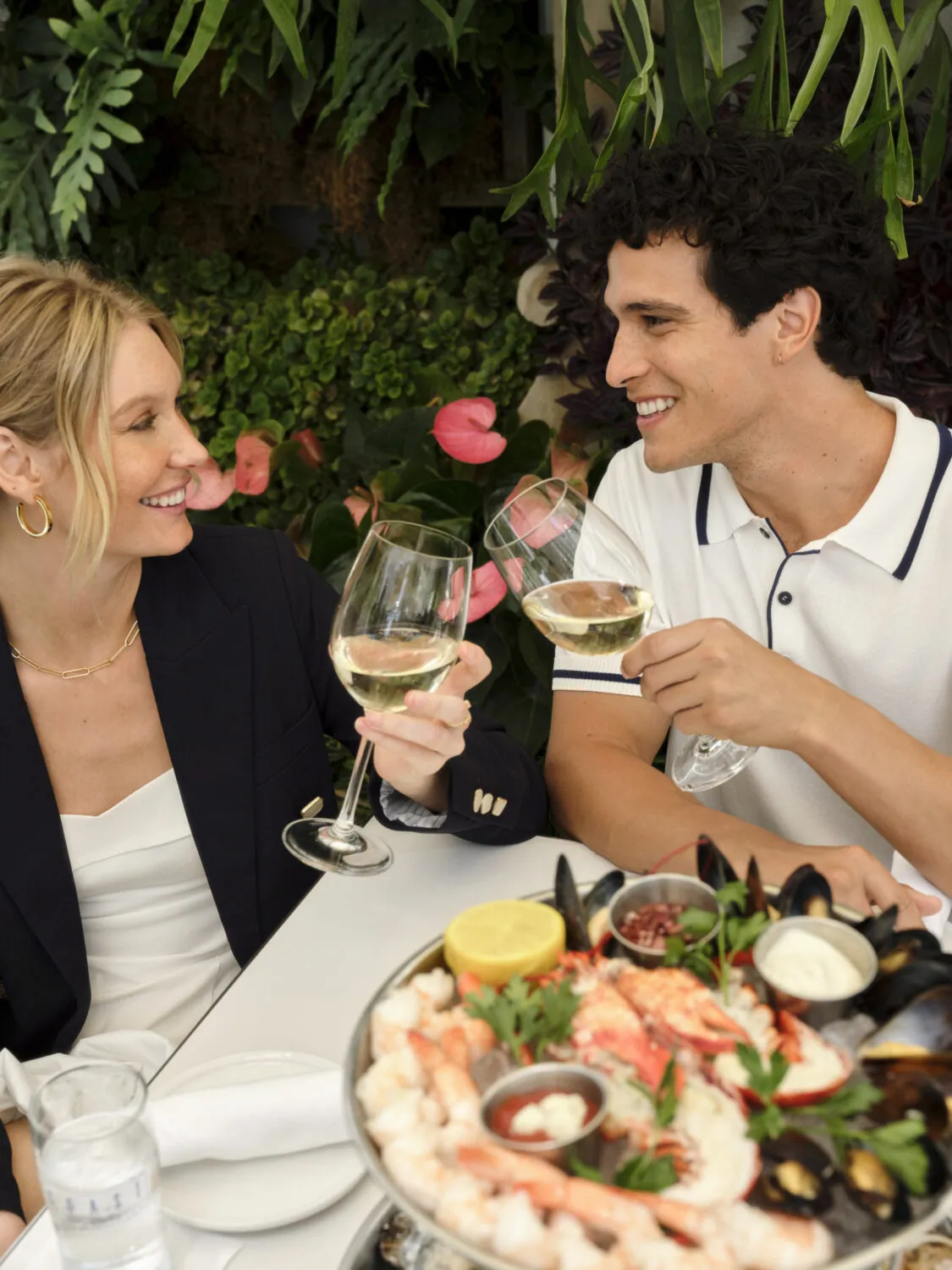 A couple toasting with wine glasses at a table with a seafood platter, including shrimp and mussels.