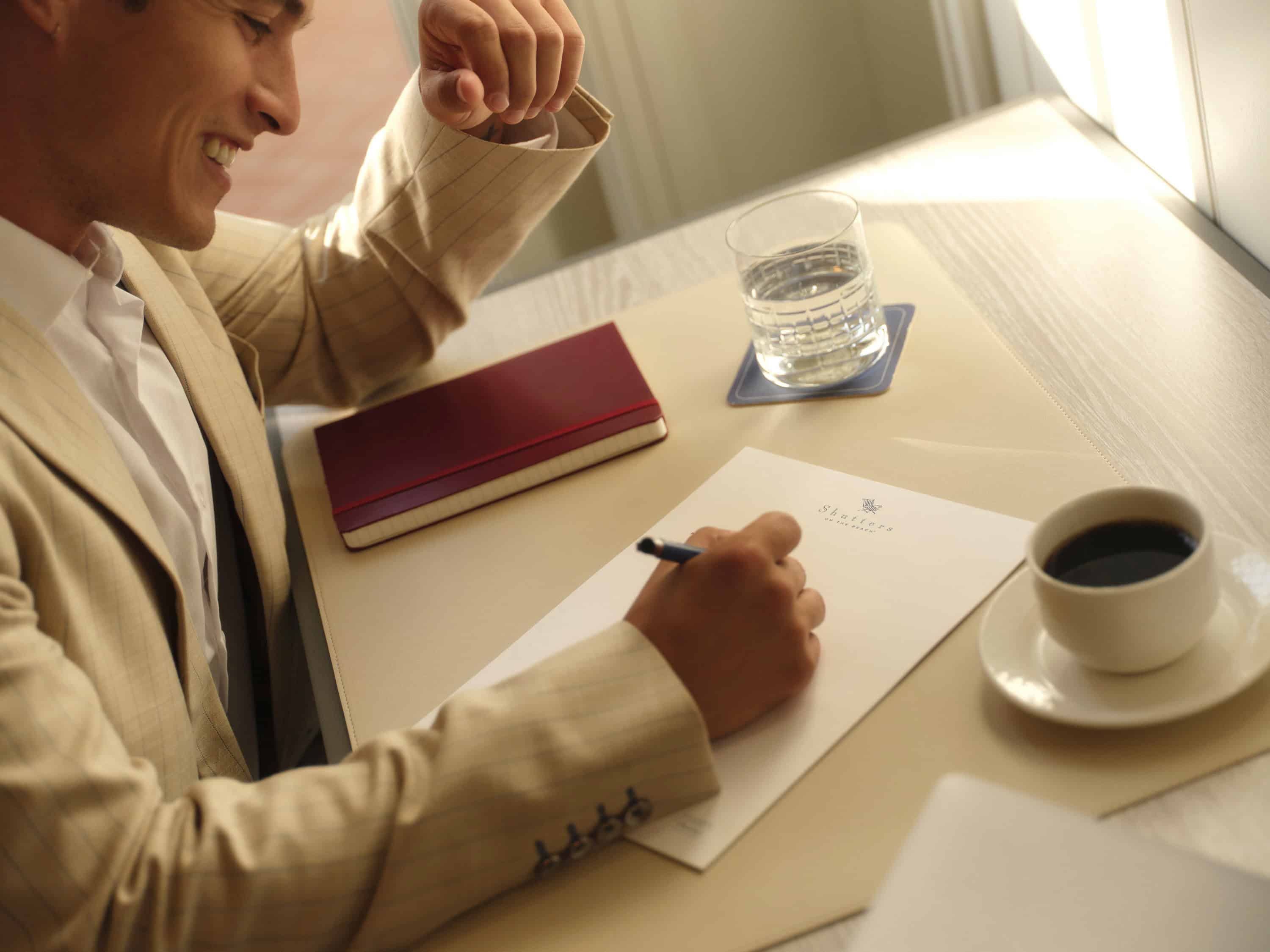 A man wearing a suit writing on a piece of paper at a table, with a cup of coffee, and a glass of water.