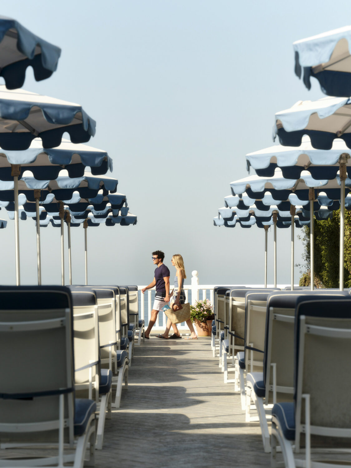 A couple walks among rows of beach umbrellas and lounge chairs by the water.