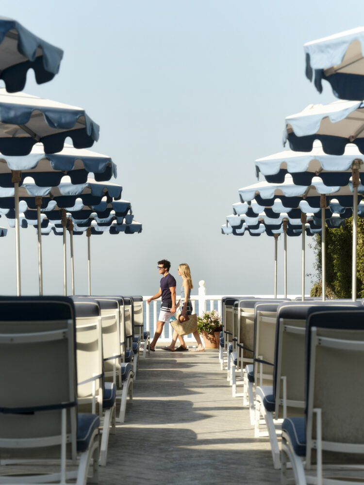 A couple walks among rows of beach umbrellas and lounge chairs by the water.