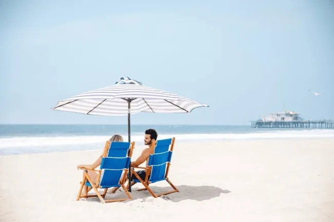 Couple relaxing on blue beach chairs under an umbrella, facing the ocean with a pier in the distance