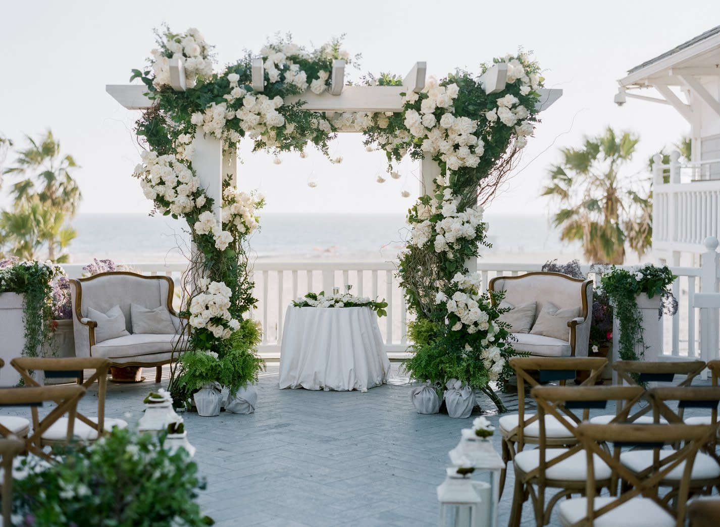 A floral wedding arch by the ocean, covered in white roses and greenery, with a beachside view.