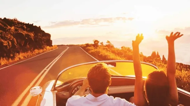 Couple driving a convertible on an open road at sunset, with arms raised and golden sunlight.