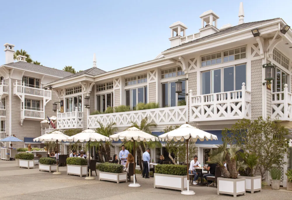 Outdoor restaurant with umbrellas, tables, patrons, and a coastal-style building.