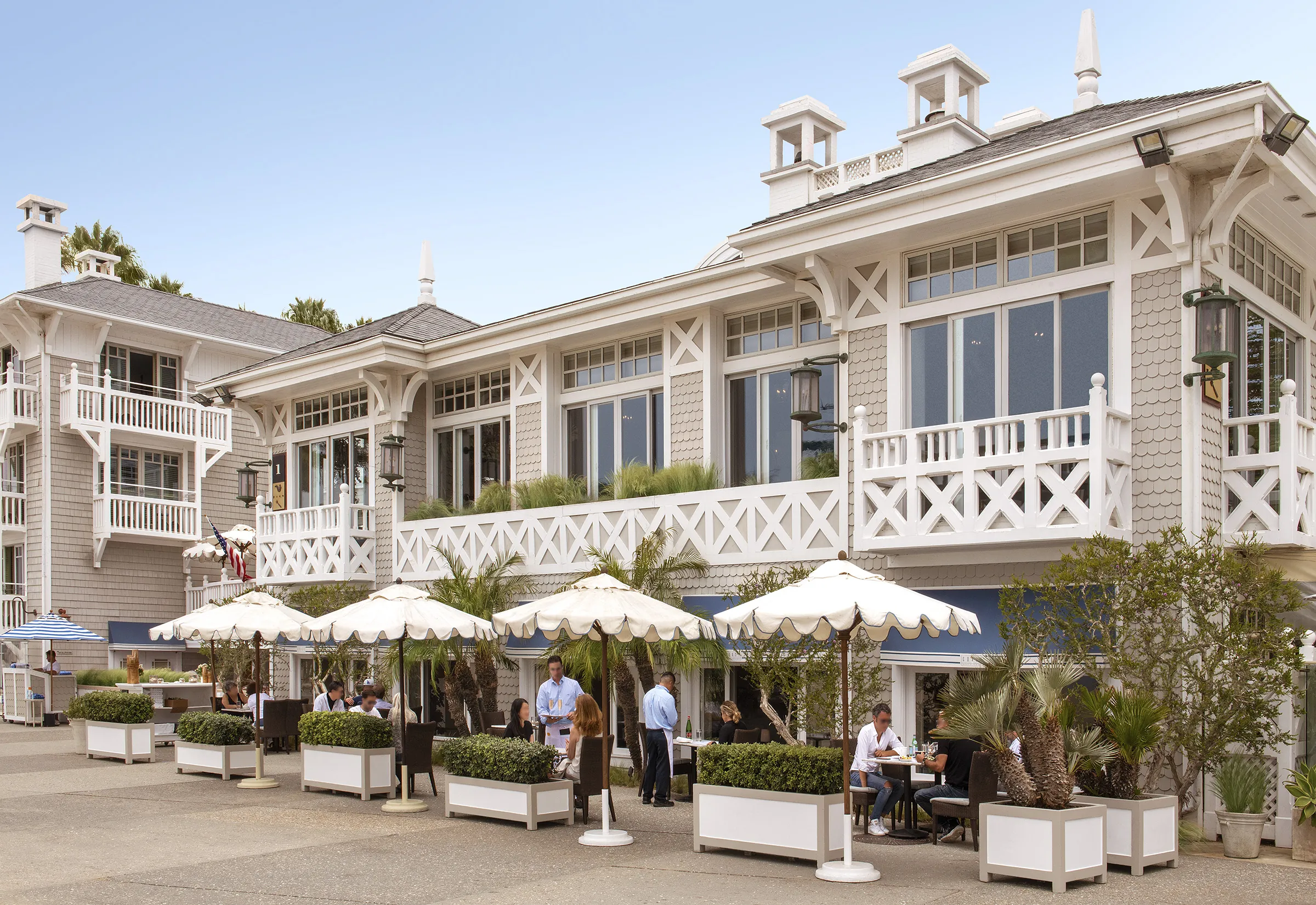 Outdoor restaurant with umbrellas, tables, patrons, and a coastal-style building.