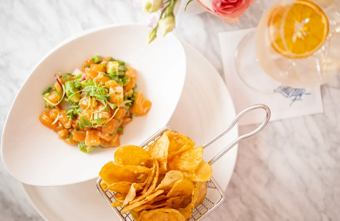 A bowl of salmon tartare with avocado, herbs, and a side of crispy chips and a cocktail.
