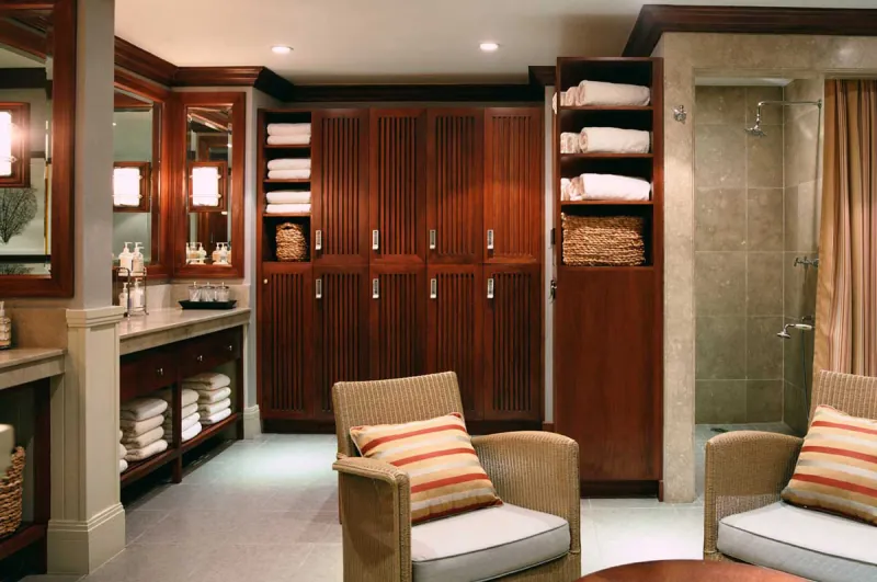 Elegant spa locker room with wooden cabinets, plush seating, stacked towels, and a tiled shower.