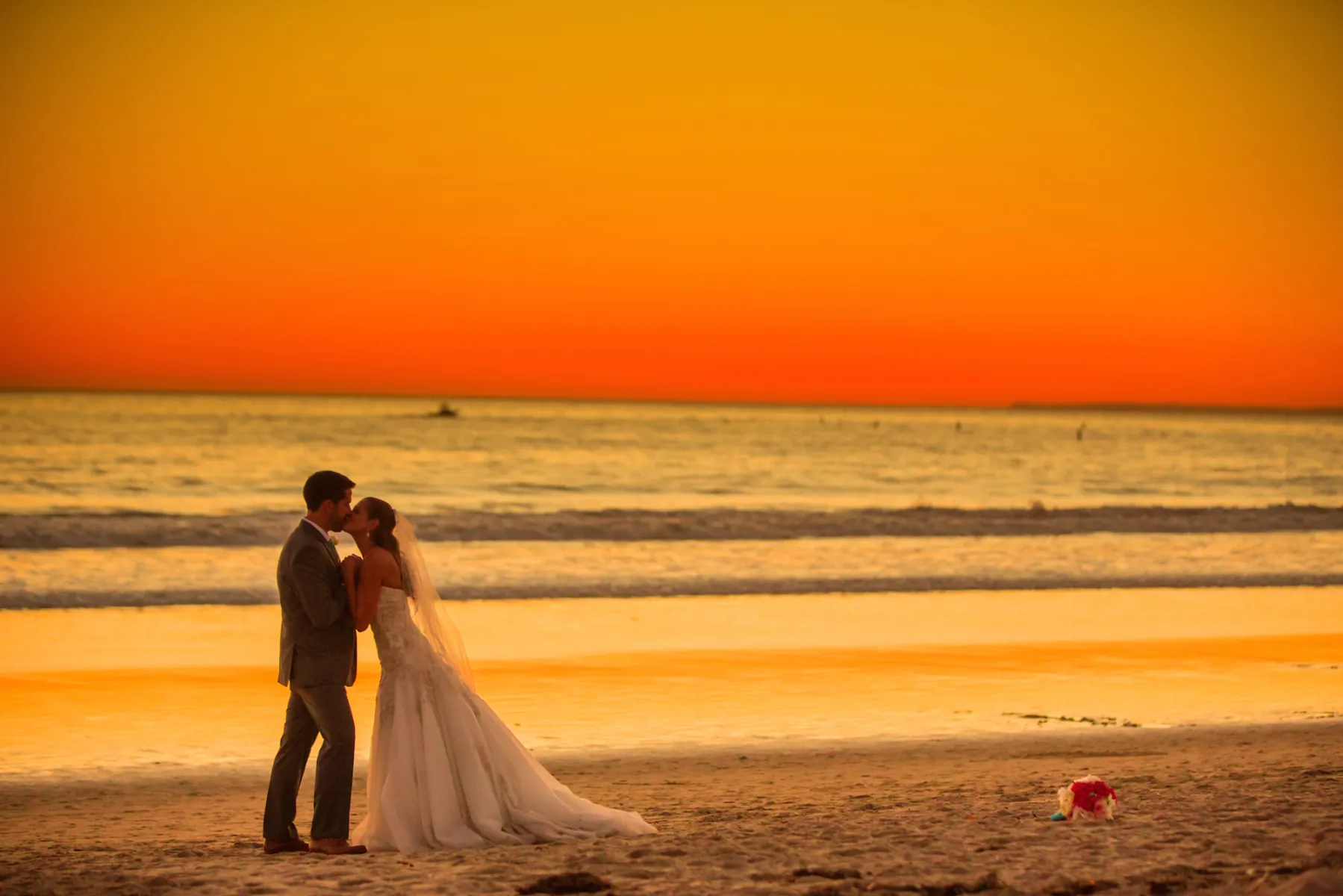 A couple in wedding attire kissing on a beach at sunset with the ocean and vibrant sky behind them.