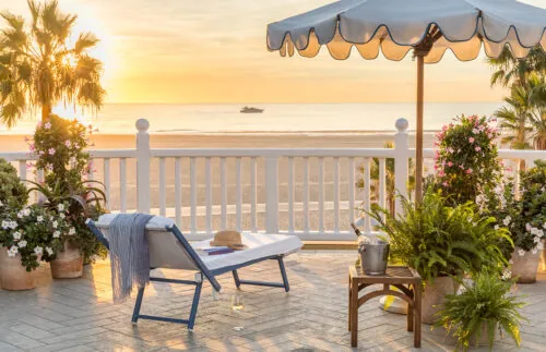 A relaxing beachside scene with a lounge chair, plants, and a glass of wine at sunset.