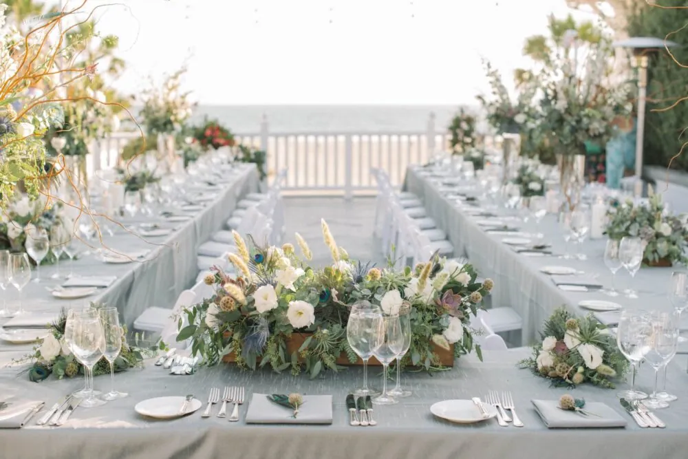 Elegant outdoor wedding reception setup with long tables, floral centerpieces, and ocean views.