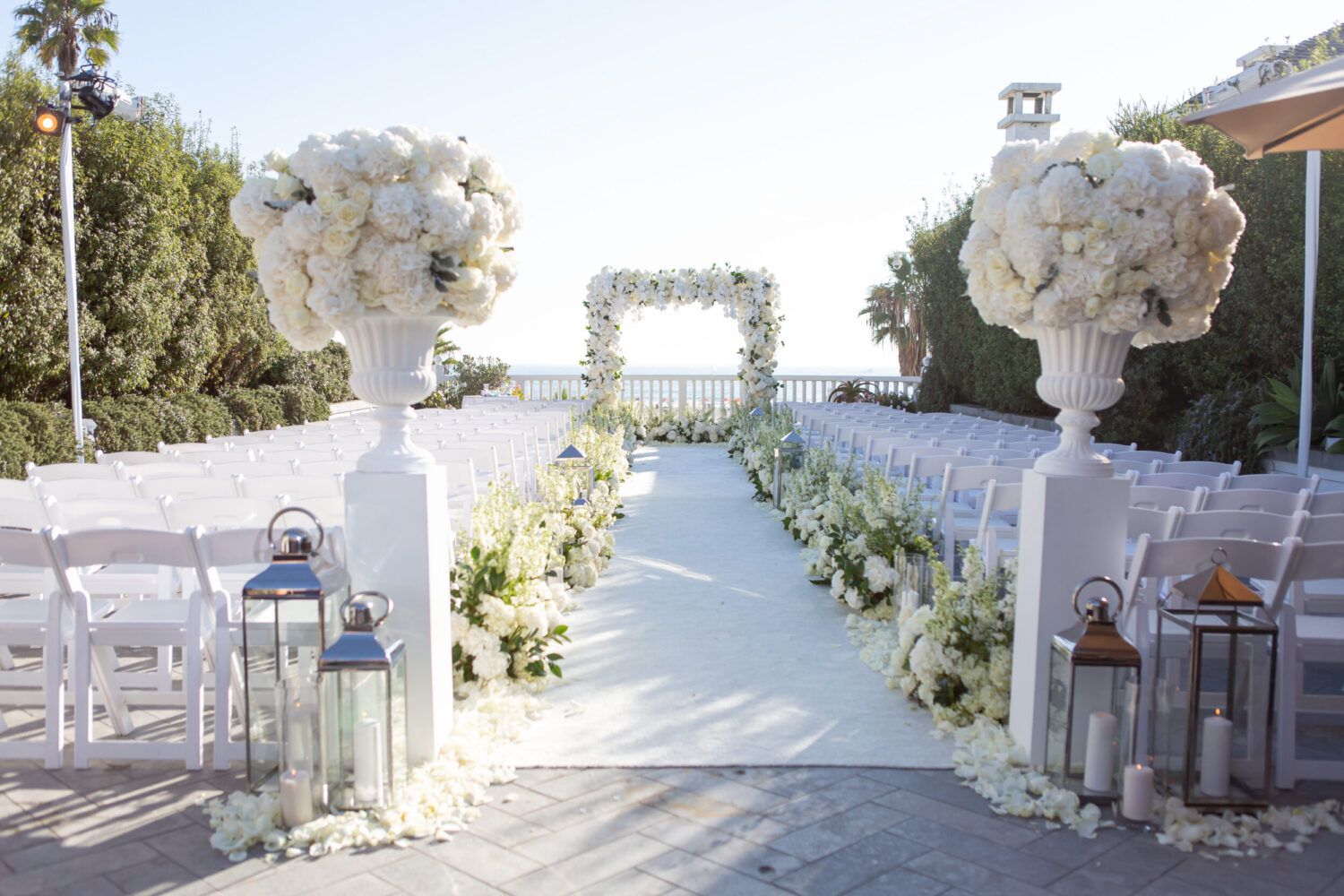 Elegant wedding ceremony setup with white floral arrangements, lanterns, and ocean view.