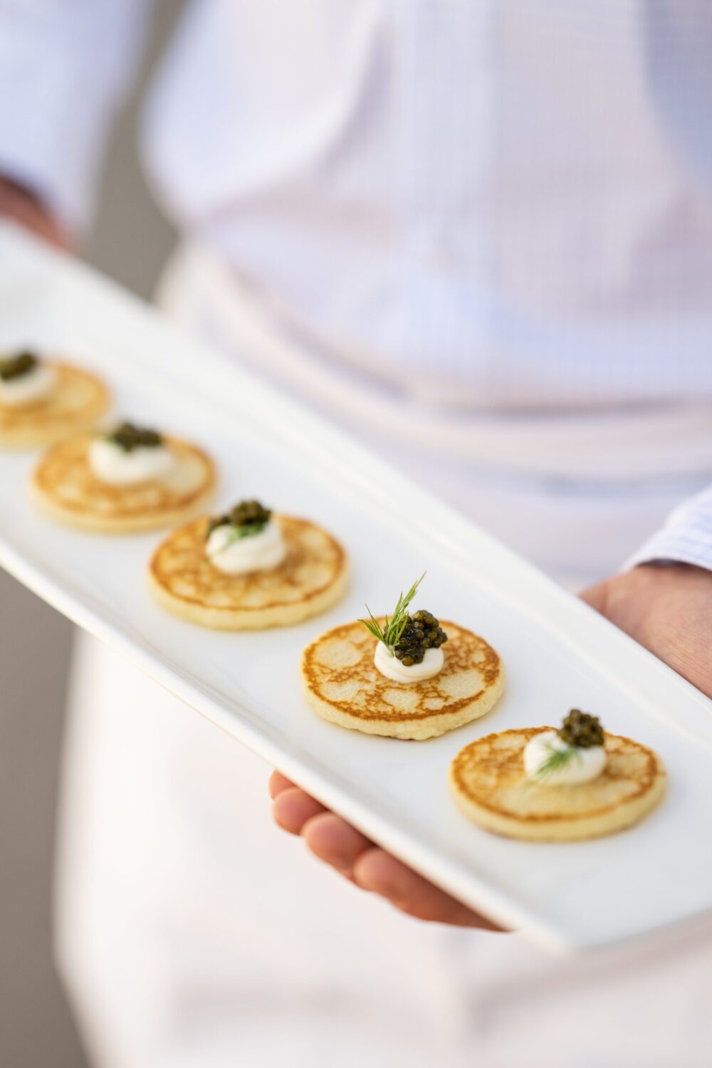 A waiter serving blinis topped with caviar and sour cream on a white plate.