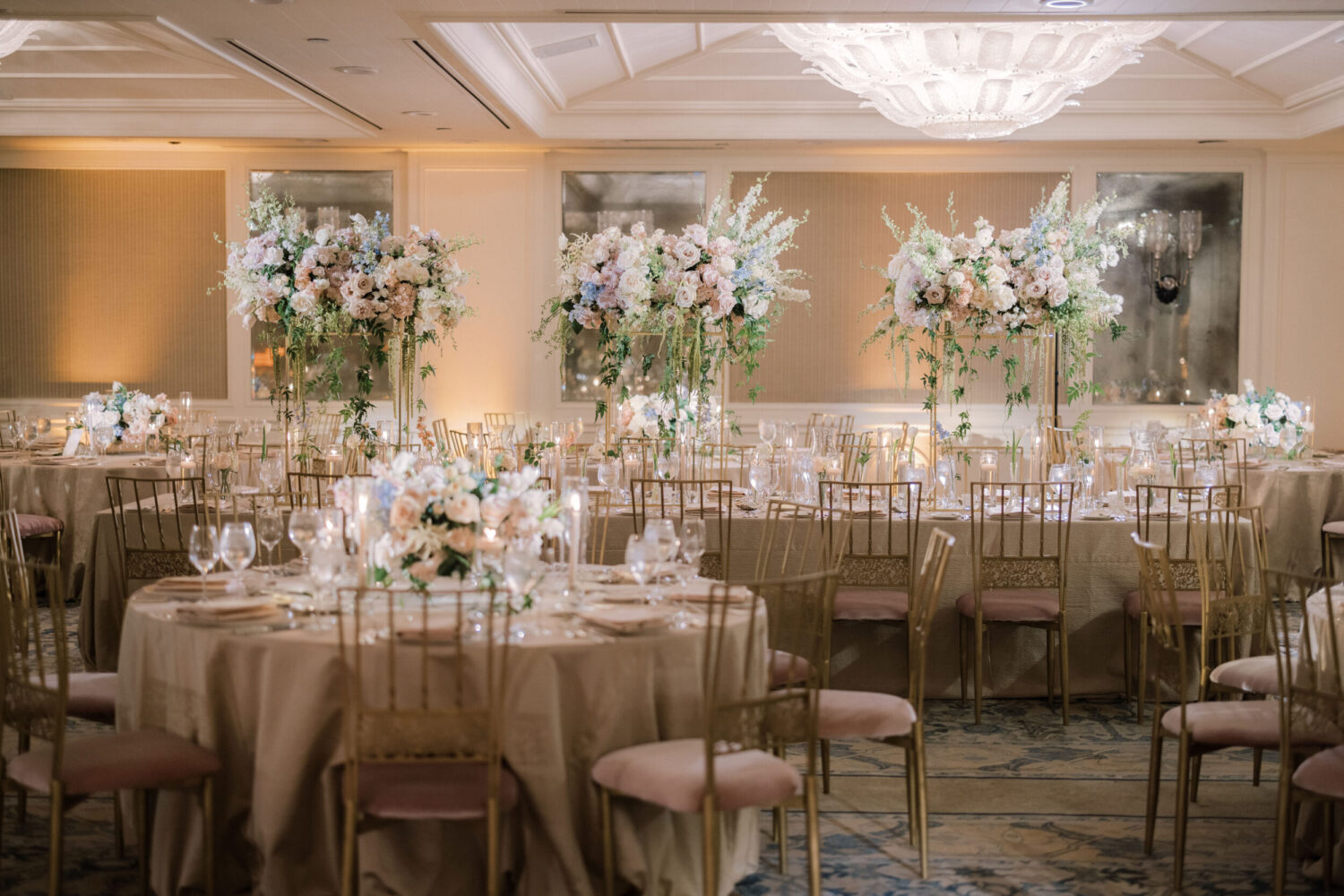 Wedding reception with floral decor, gold chairs, candles, and a grand chandelier.