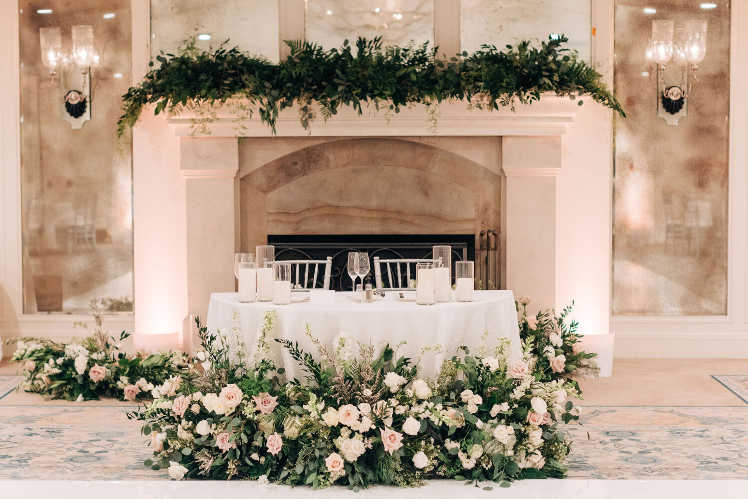 Romantic sweetheart table with floral arrangements and candlelit elegance by the fireplace.