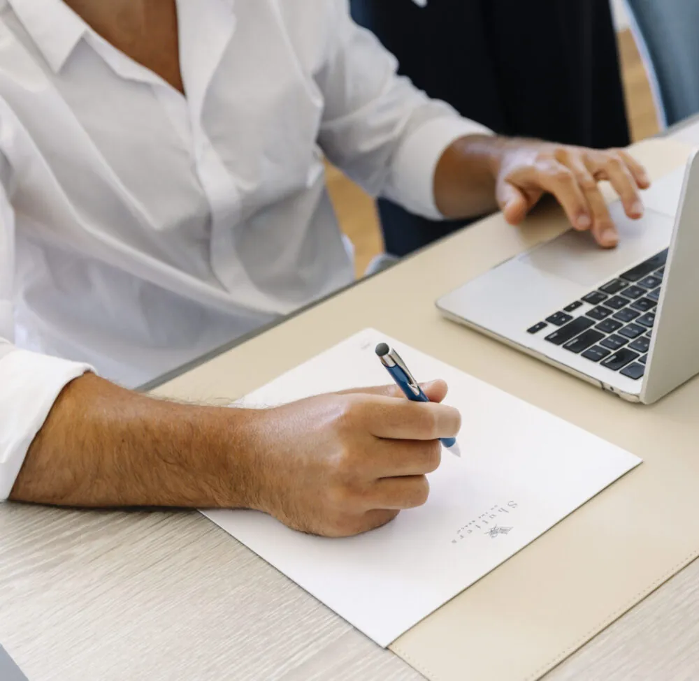 A person is writing on paper with a pen, while using a laptop on the desk.