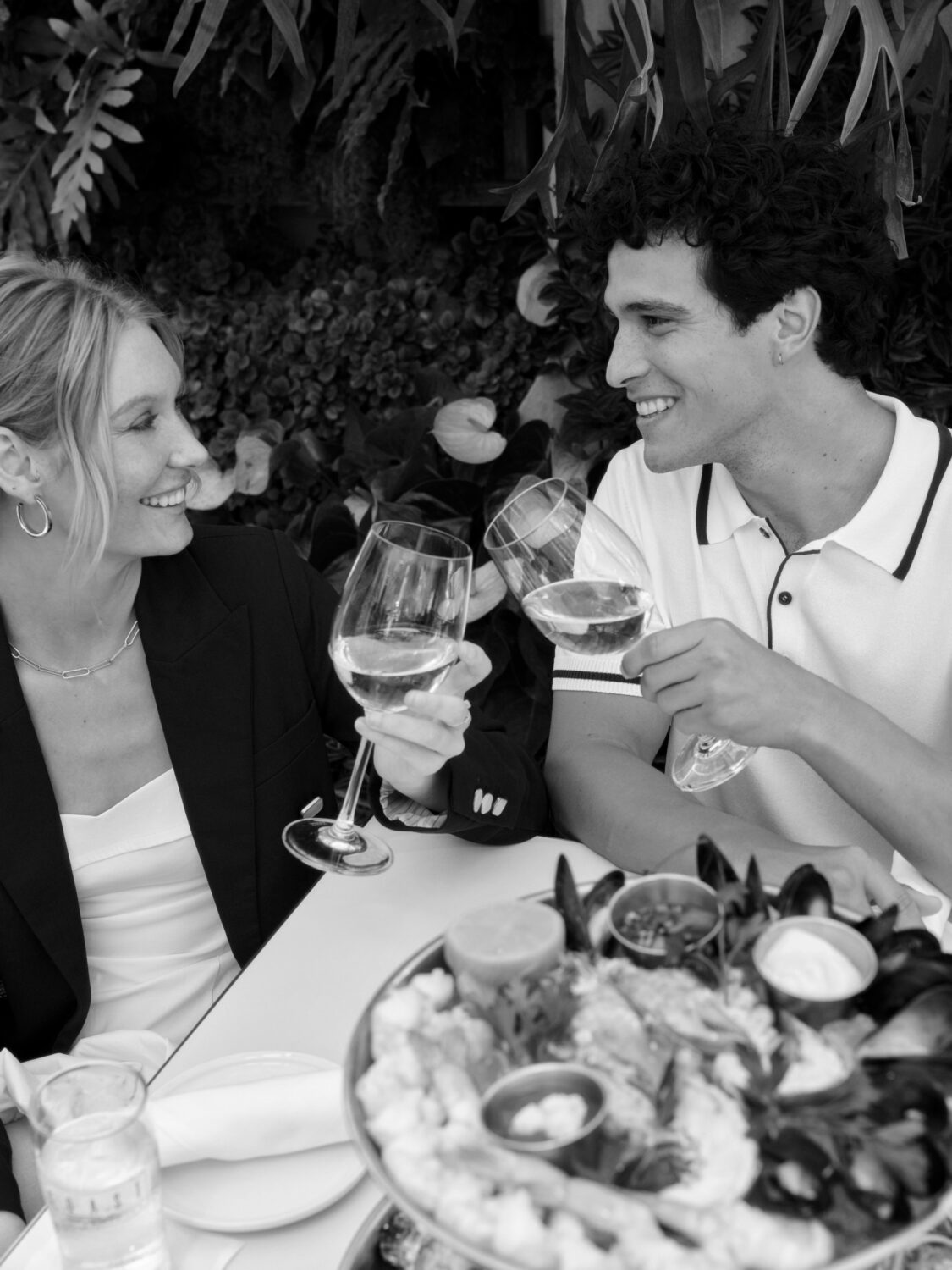 A couple smiling at each other while holding wine glasses, with a seafood platter on the table.