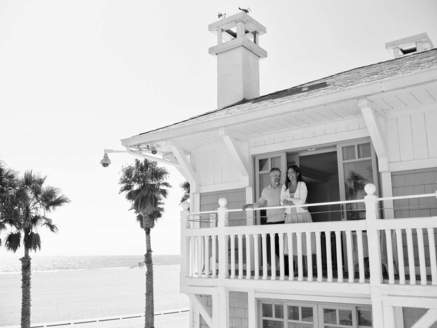 A couple standing on the balcony of a white beach house, with the ocean in the background.