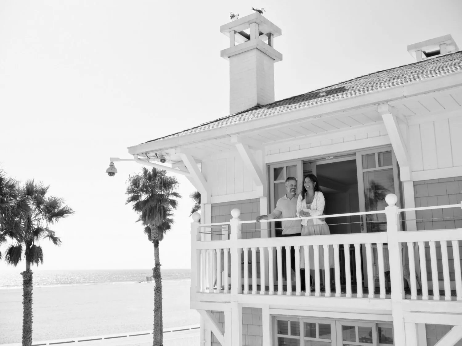 A couple standing on the balcony of a white beach house, with the ocean in the background.