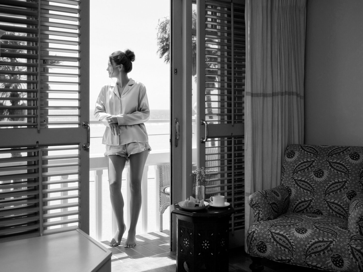 Woman in a robe standing barefoot on a balcony with shutters open, looking out at the ocean view.