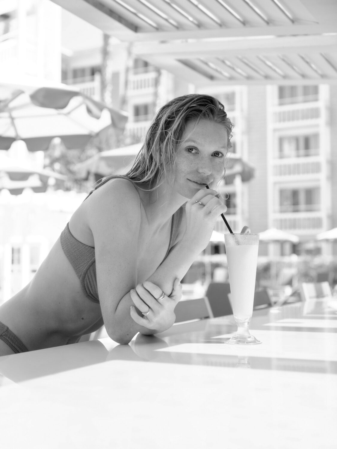 Woman in a swimsuit leaning on a table, sipping a drink through a straw at a poolside bar.