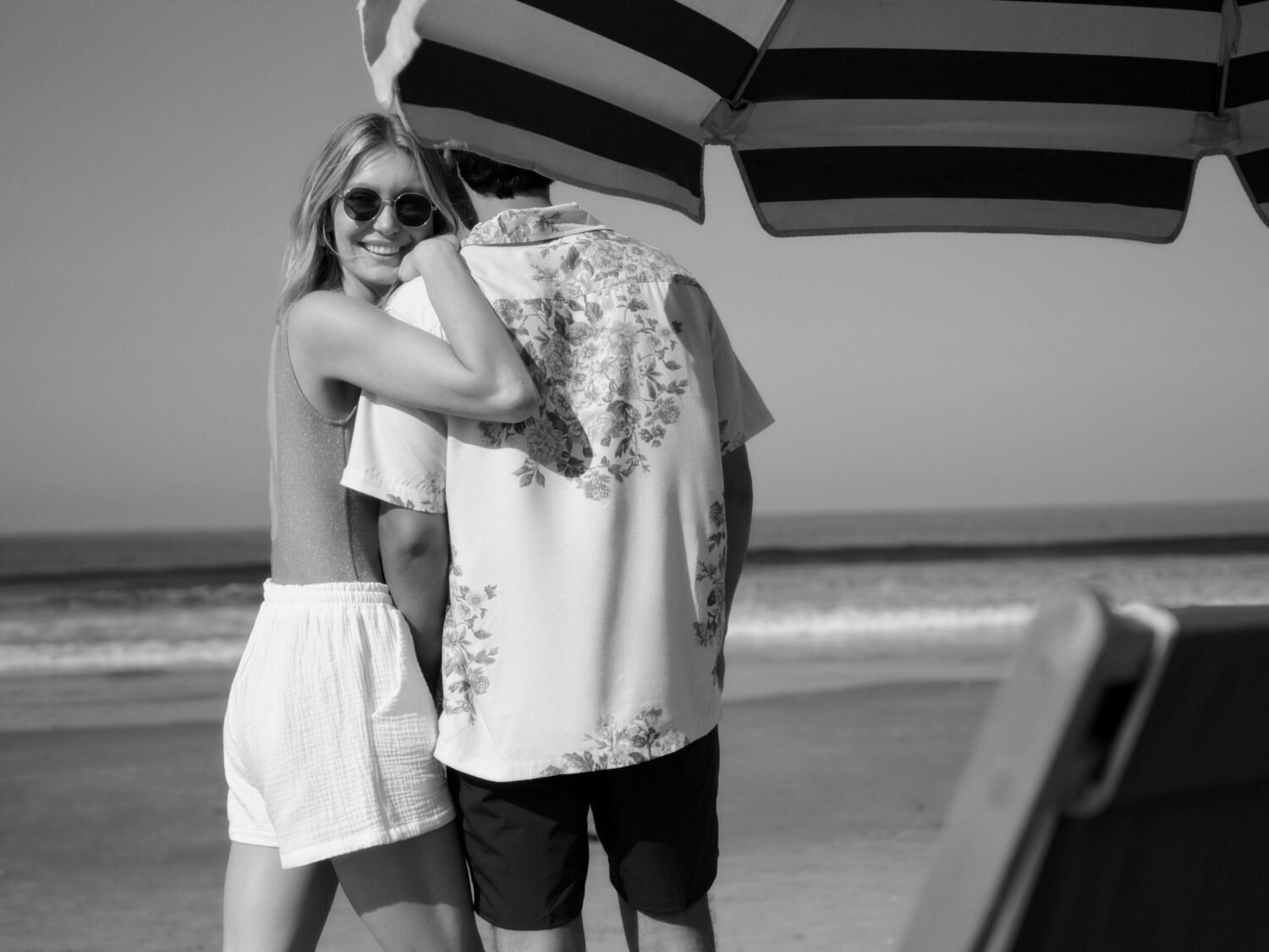 Couple embracing on the beach under a striped umbrella, with the ocean in the background.