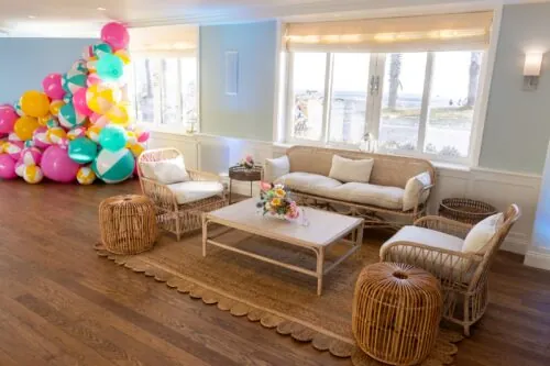 Coastal lounge with rattan furniture, cushions, ocean views, and colorful balloons.