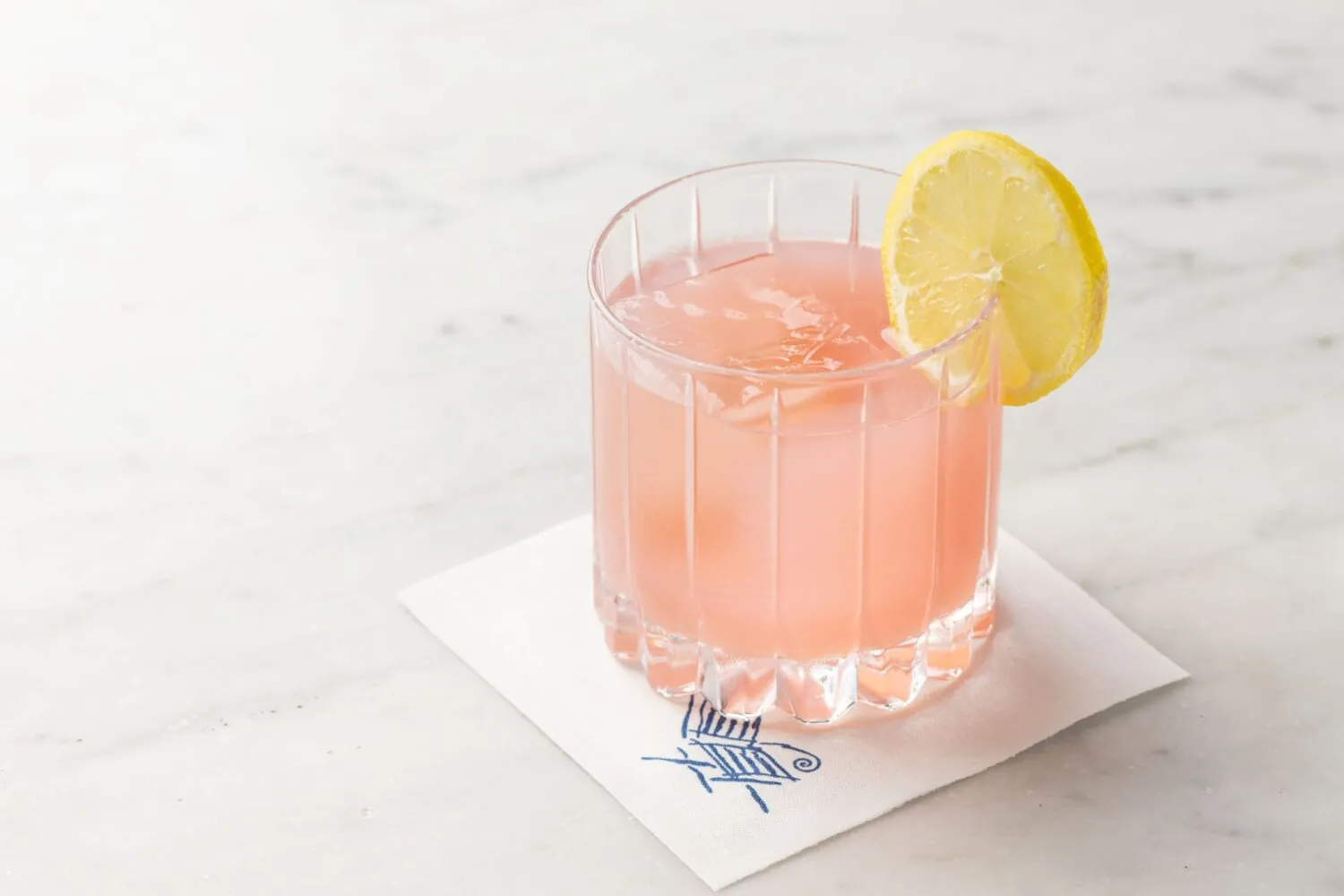A pink drink with a slice of lemon served over ice in a glass on a napkin.