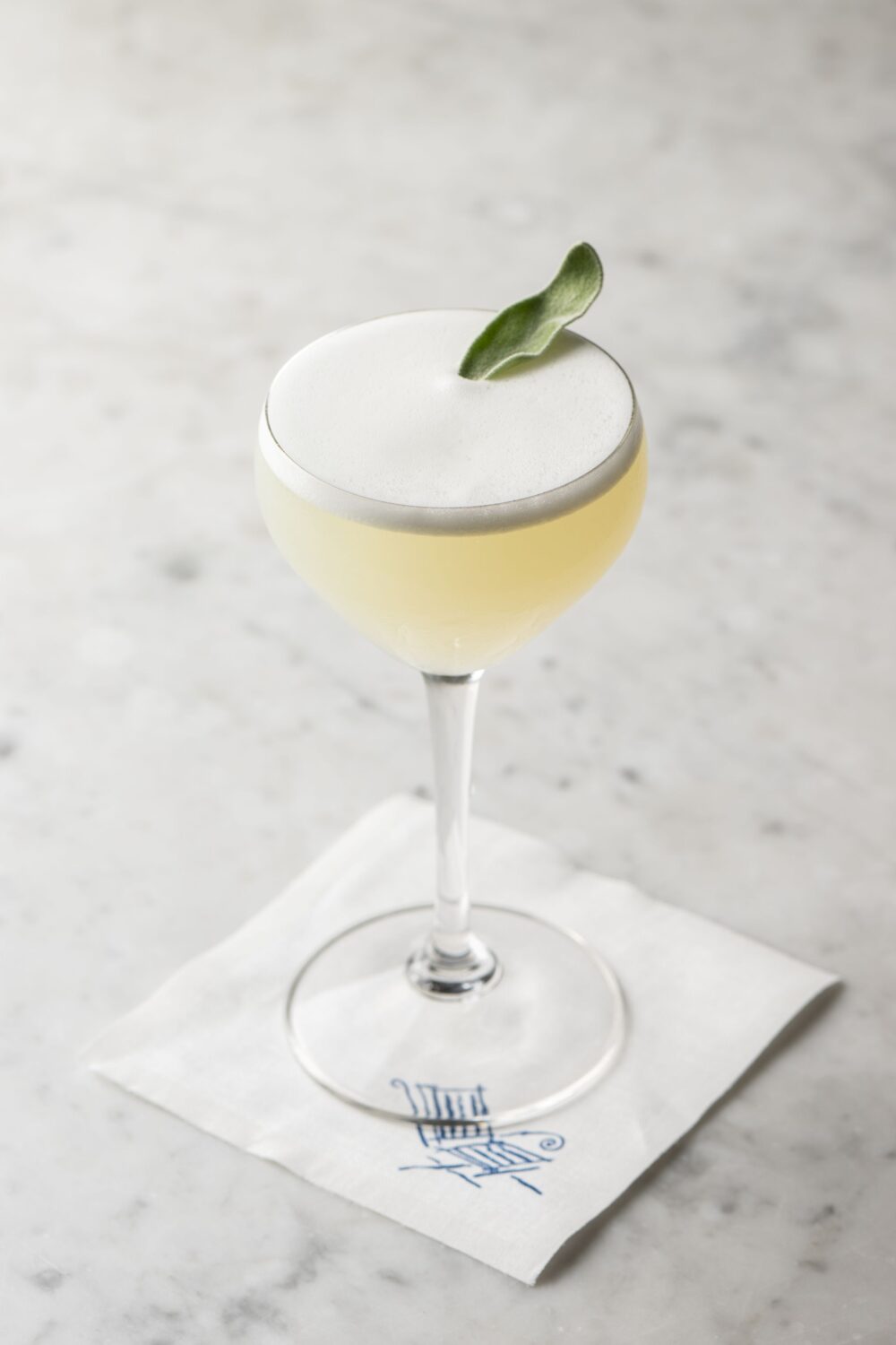 Close-up of a cocktail glass with a frothy top and sage leaf garnish on a napkin.