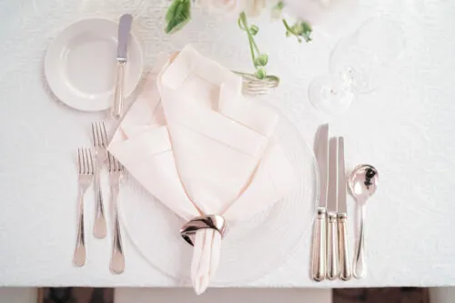 Refined table setting with silverware, a folded napkin in a ring, and a clear plate on white linen.