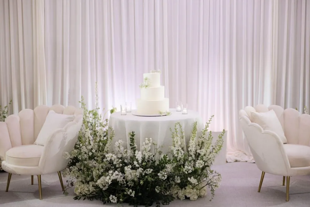 White wedding cake with delicate flowers, surrounded by floral arrangements and elegant seating.
