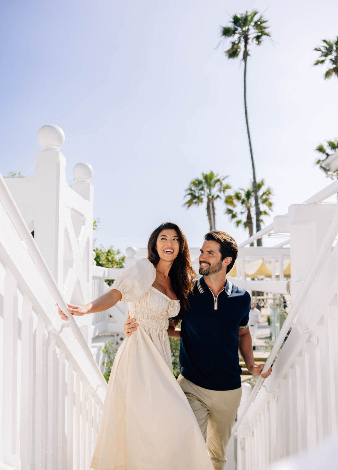A couple smiling, walking up stairs in a sunny, palm tree-filled outdoor setting.