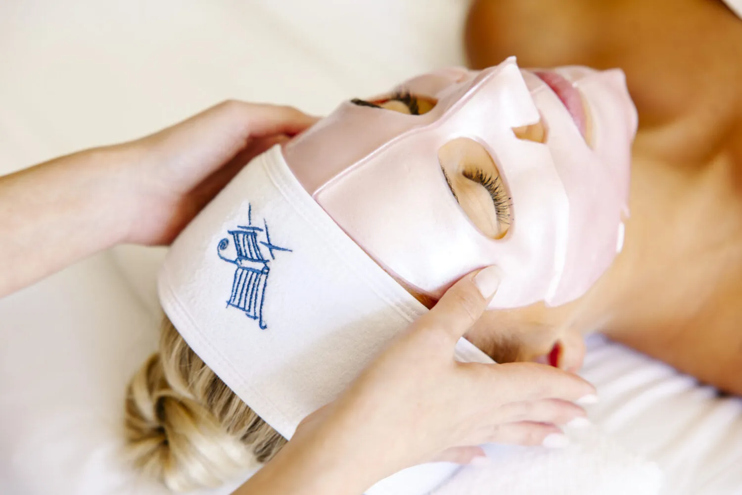 Woman receiving a spa treatment with a pink facial mask, wearing a branded headband.