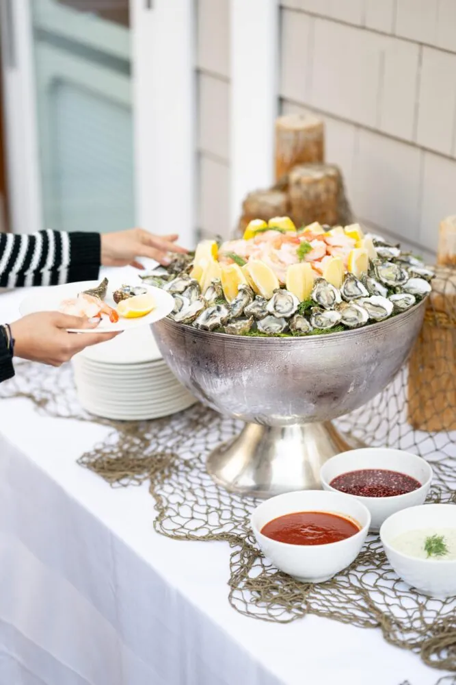 Seafood display with oysters, shrimp, lemon wedges, and sauces, as a guest fills their plate.