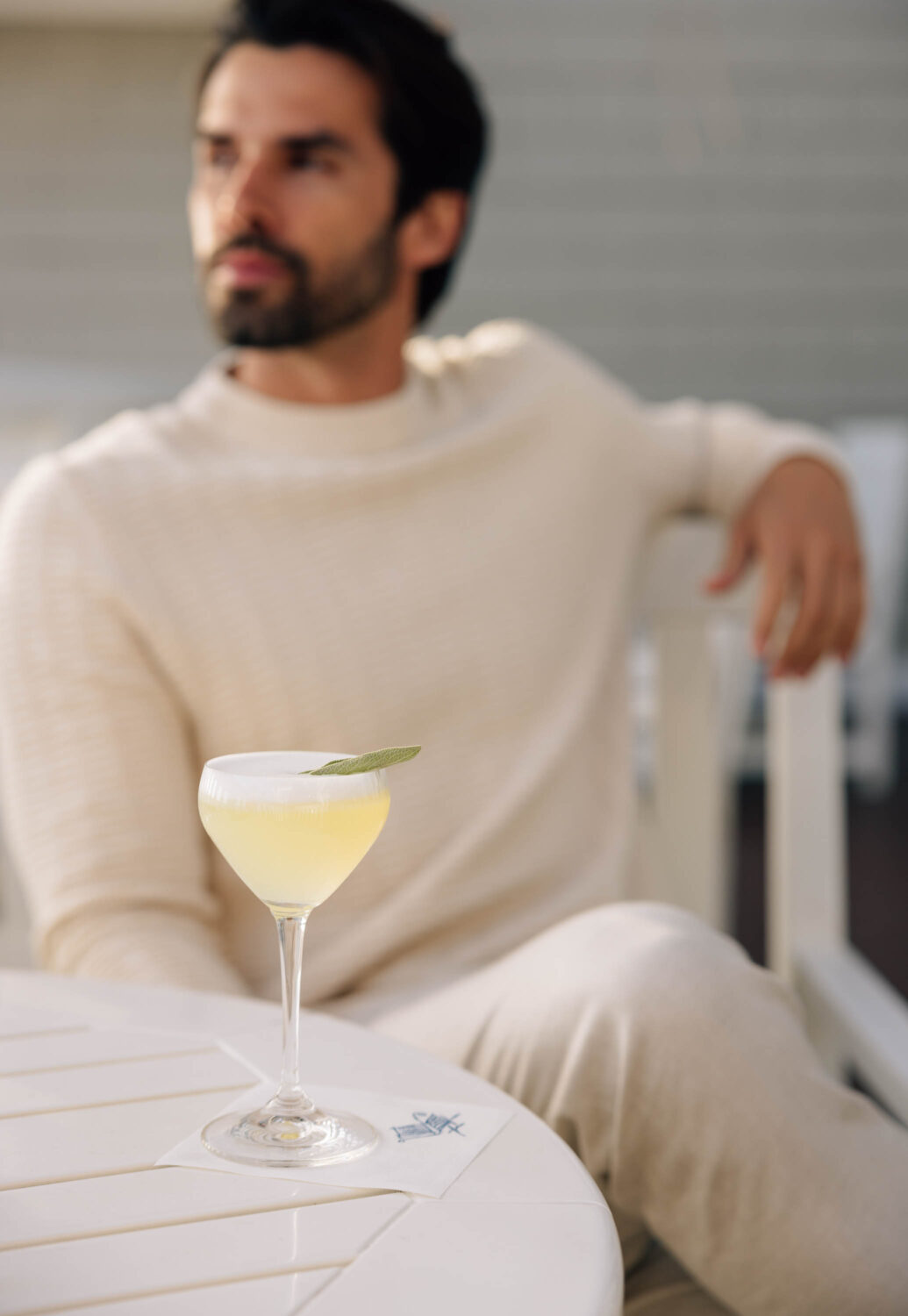 Yellow cocktail with sage on a white table, a relaxed man in a cream sweater in the background.
