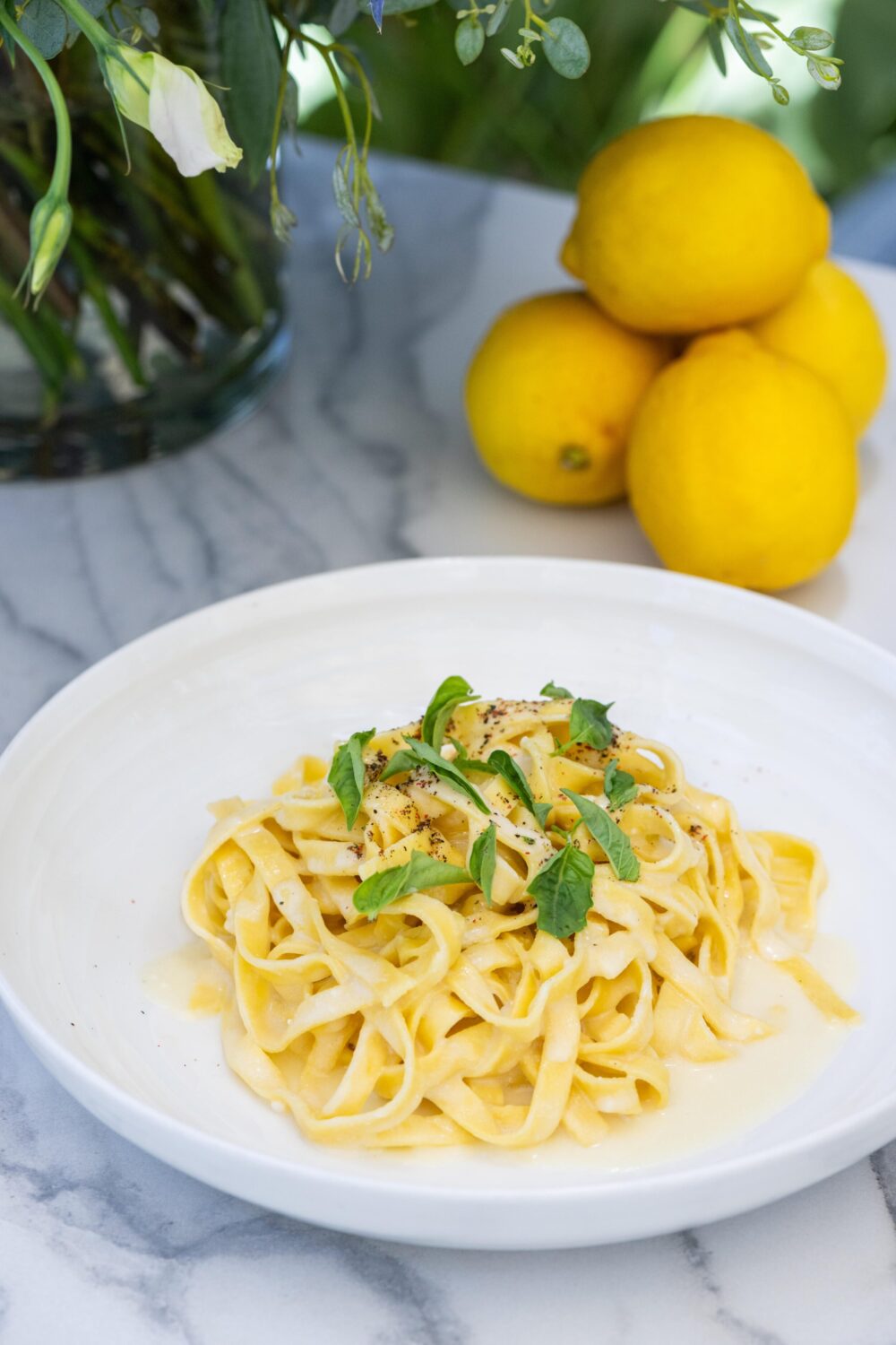 A plate of fettuccine pasta with lemon, basil, and black pepper, with lemons in the background.